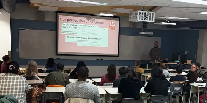 Une salle de cours vue du fond : le public, assis aux tables, fait face à une diapositive projetée et à la personne qui présente.